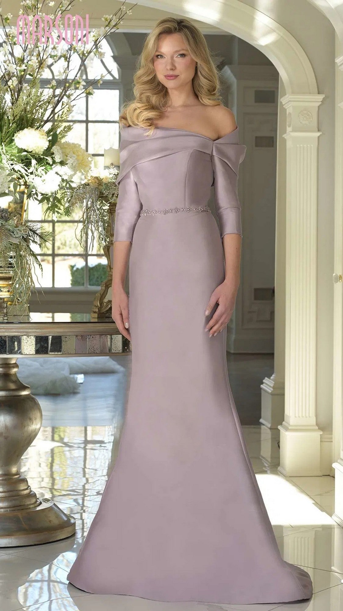 A woman stands indoors wearing the Marsoni MV1375 long formal dress in dusty rose, featuring a floor-length skirt, off-shoulder neckline, three-quarter sleeves, and a jeweled belt.